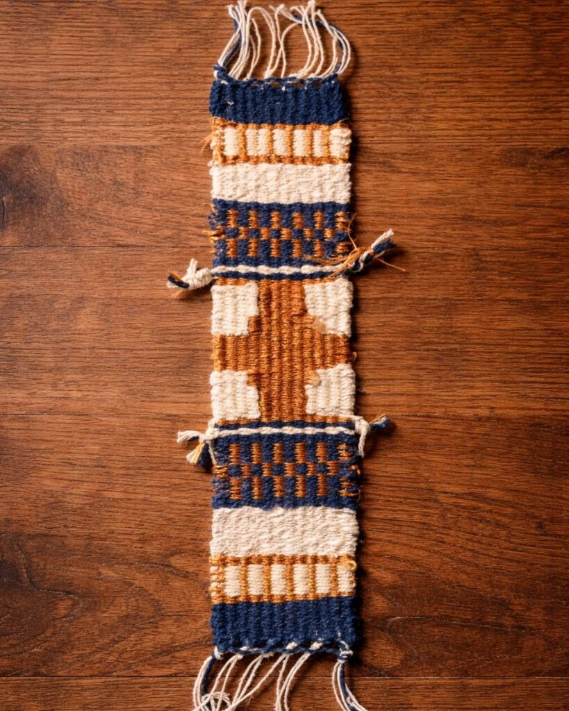 Detailaufnahme eines handgewebten Lesezeichens von Wiedengold mit abstraktem Muster in Gold- und Naturtönen.