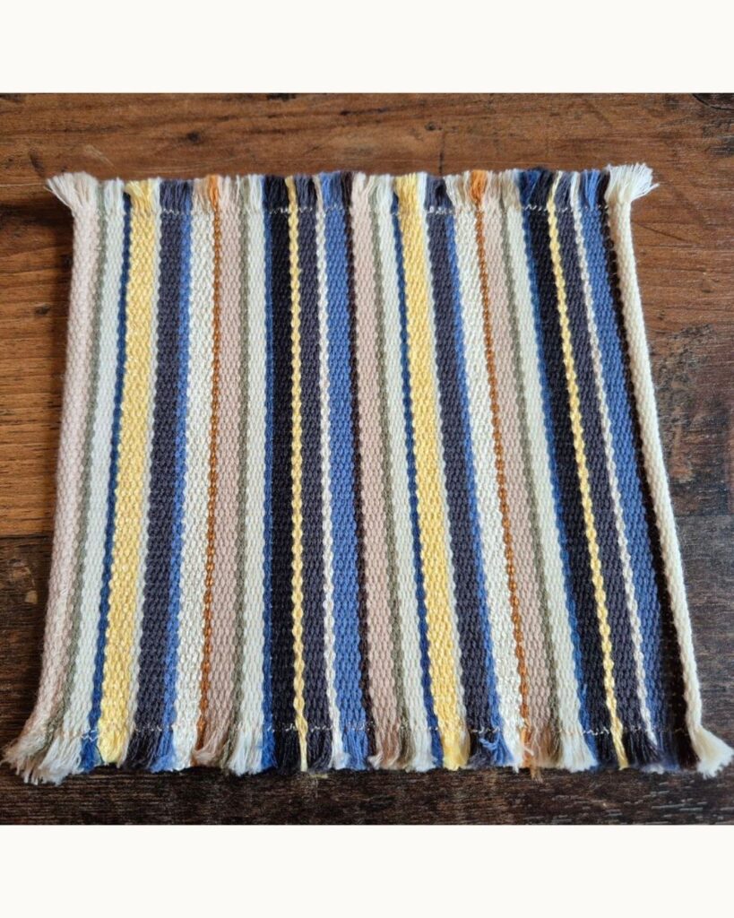 Handgewebter Untersetzer aus Naturmaterialien mit Streifen in Blau, Gelb und weiteren sommerlichen Farben.