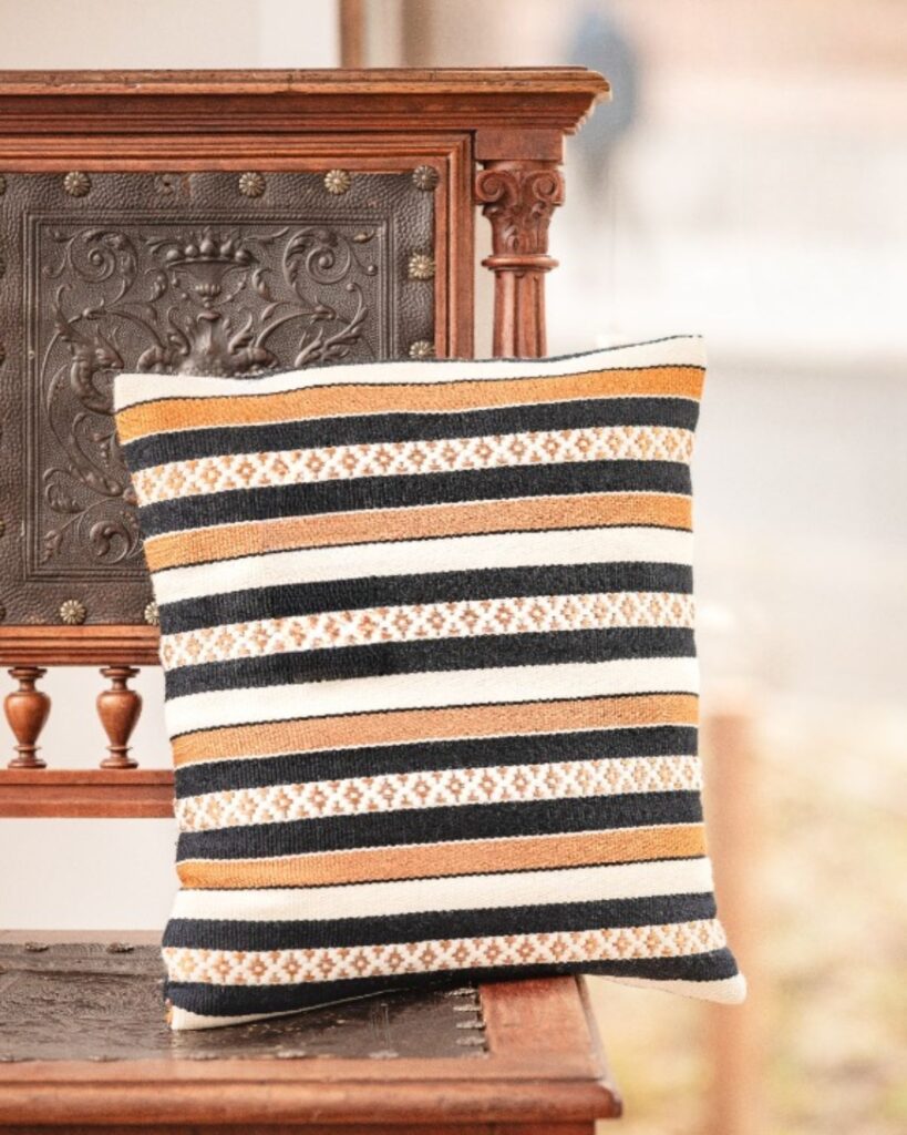 Handgewebtes Kissen in Wolle und Viskose mit grafischen Streifen in Gold, Schwarz und Weiß, auf antikem Sessel.