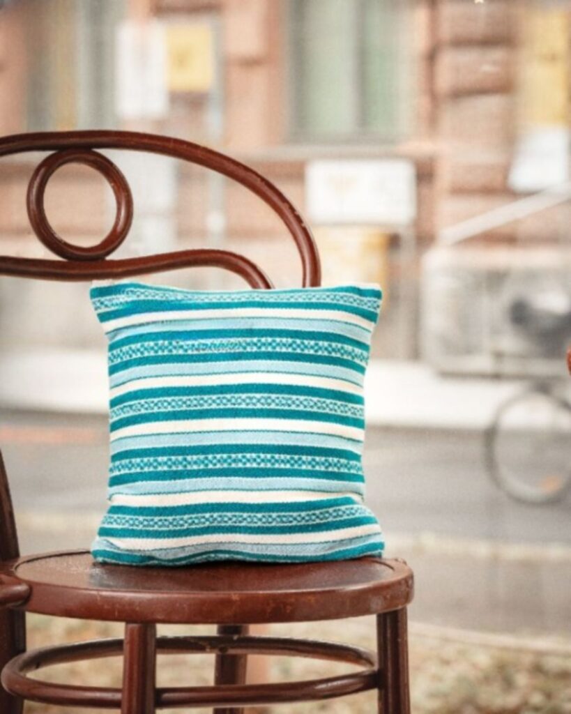Handgewebtes Kissen von Wiedengold mit Streifen in Türkis und Weiß, auf einem Stuhl präsentiert.