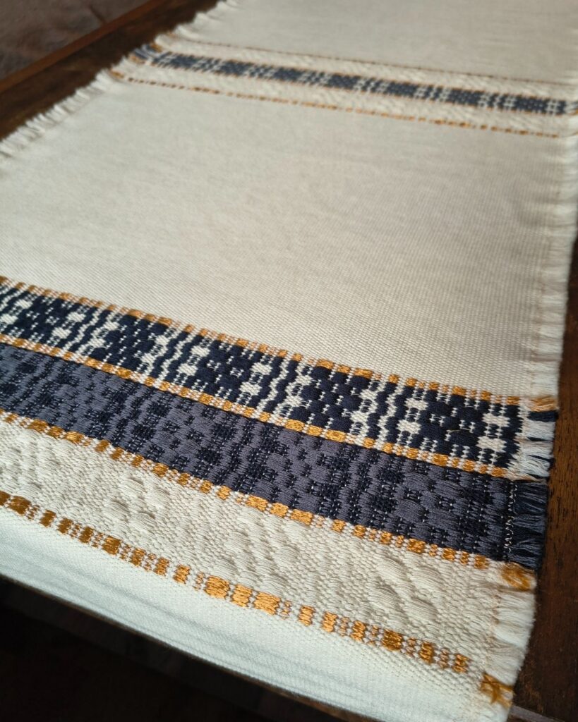Detailaufnahme eines handgewebten Tischläufers aus Wolle und Viskose mit dunkelblauer Ornamentborte und goldfarbenen Akzenten.