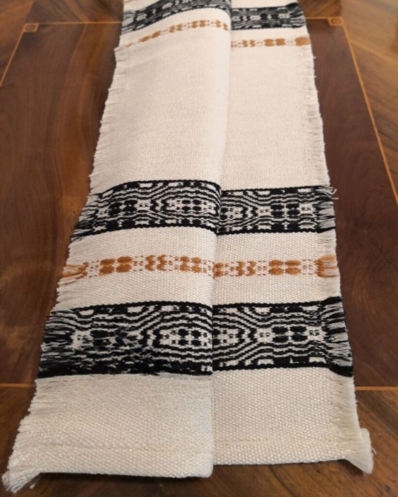 Detailaufnahme eines handgewebten Tischläufers aus Wolle und Viskose von Wiedengold mit schwarz-goldener Ornamentborte auf weißem Grund.