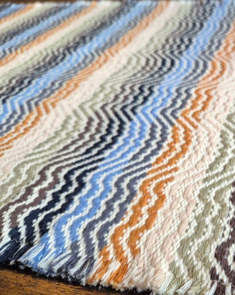 Detailansicht eines handgewebten Tischläufers in Wolle von Wiedengold mit wellenförmigen Streifen in Blau, Ocker und Rosé.