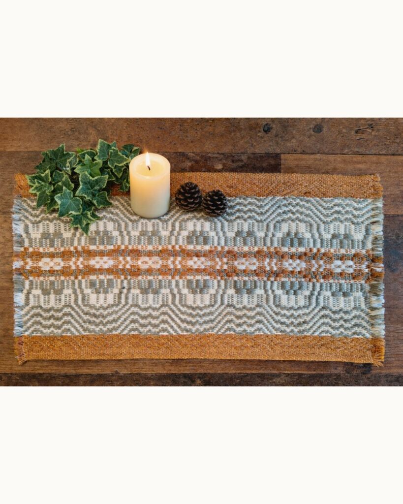 Handgewebter Tischläufer in Wolle und geometrischem Ornament in Olivgrün, Ocker und Weiß, dekoriert auf Holztisch.