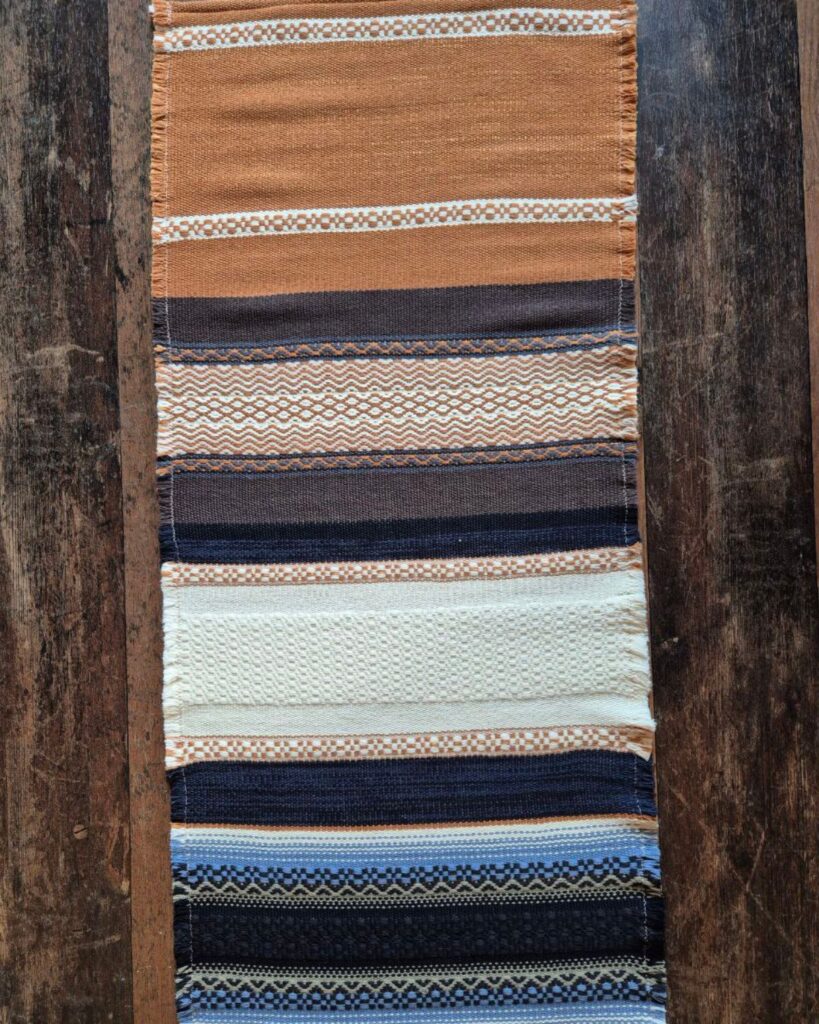 Handgewebter Tischläufer von Wiedengold in Wolle und Viskose mit Streifen in Braun, Blau, Ocker und Weiß, rhythmisch komponiert.