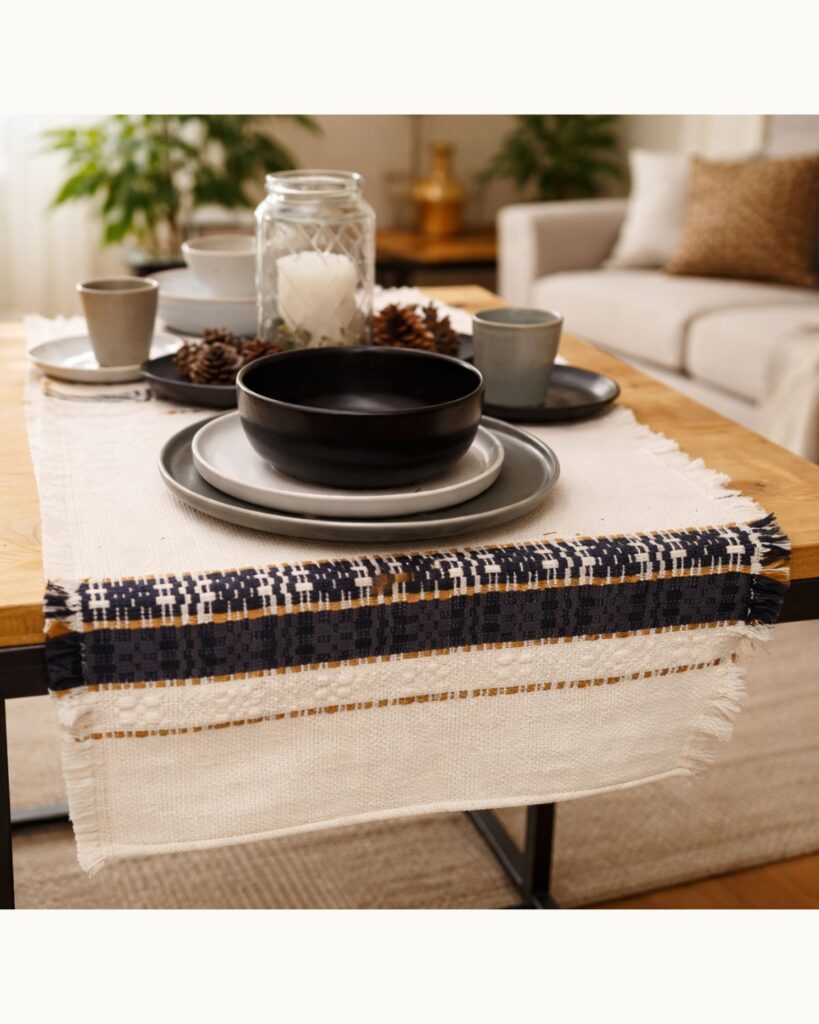 Handgewebter Tischläufer aus Wolle und Viskose in Weiß mit dunkelblauer Ornamentborte und feinen goldfarbenen Akzenten, arrangiert auf einem gedeckten Tisch.