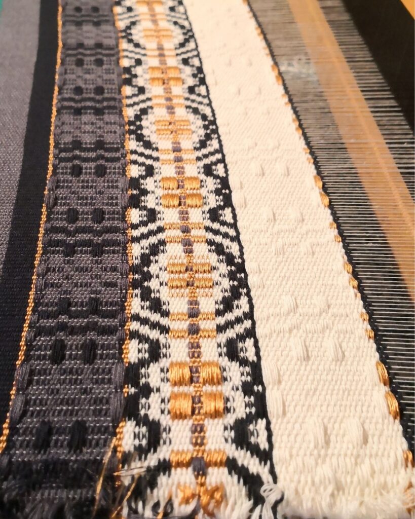Webprobe eines in Wolle handgewebten Tischläufers von Wiedengold mit dunkelblauer Fläche und goldener Ornamentborte am Webstuhl.