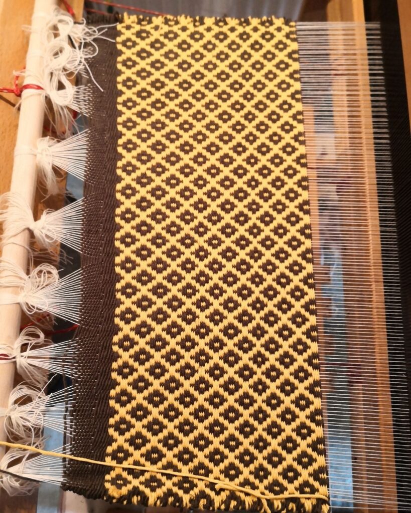 Handgewebter Stoff mit Ornamentmuster in Gelb und Schwarz am Webstuhl.