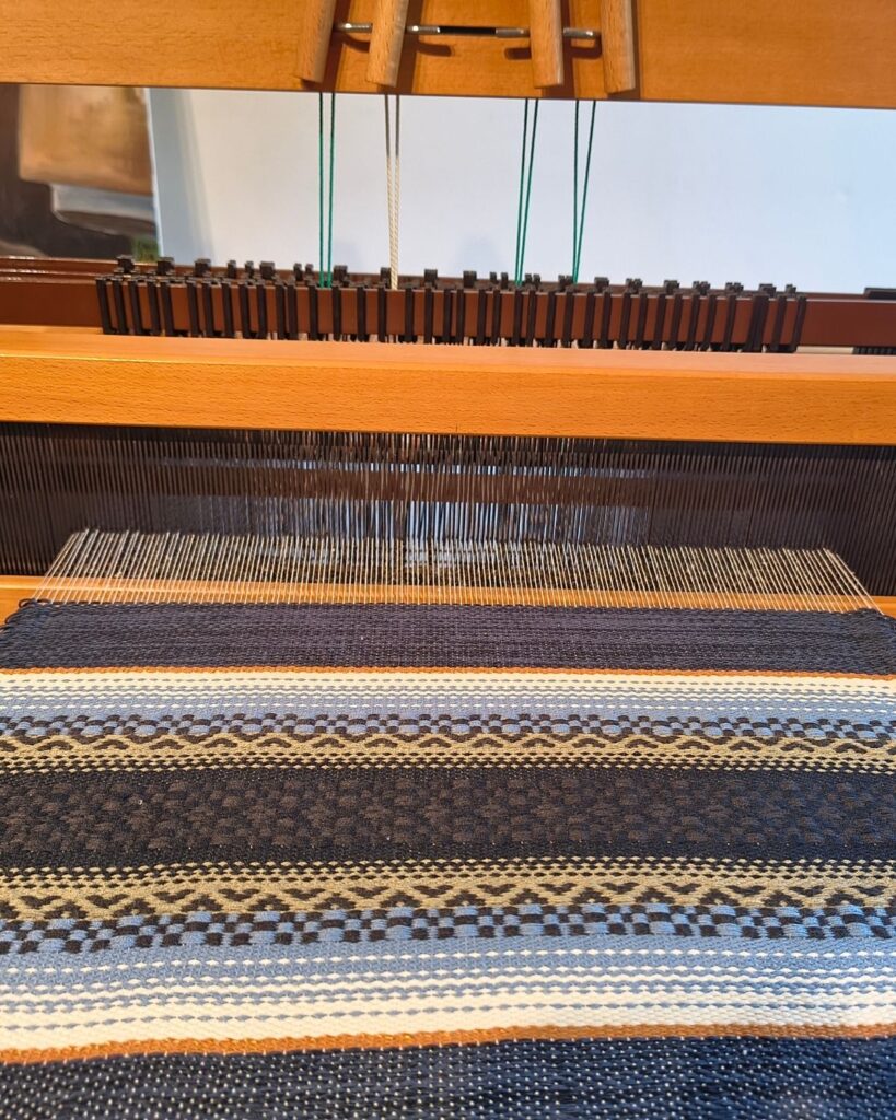 Webprobe eines handgewebten Tischläufers von Wiedengold in Wolle und Viskose mit Streifen in Braun, Blau und Weiß am Webstuhl.