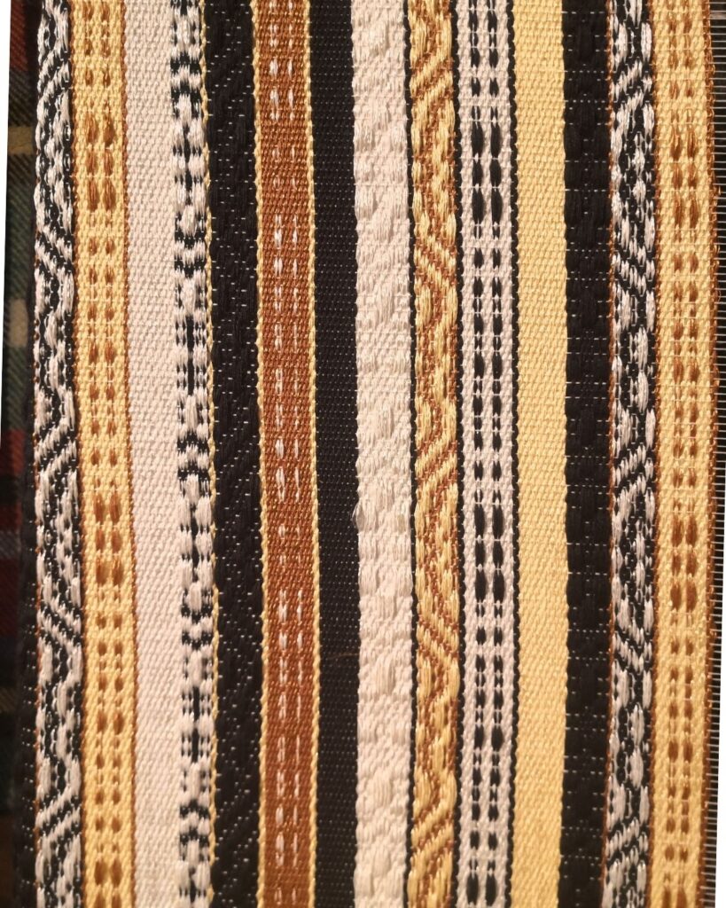 Detailansicht der Webstruktur einer handgewebten Tasche aus Naturmaterialien mit Streifen und Ornamentik in Blau, Weiß und Ocker.