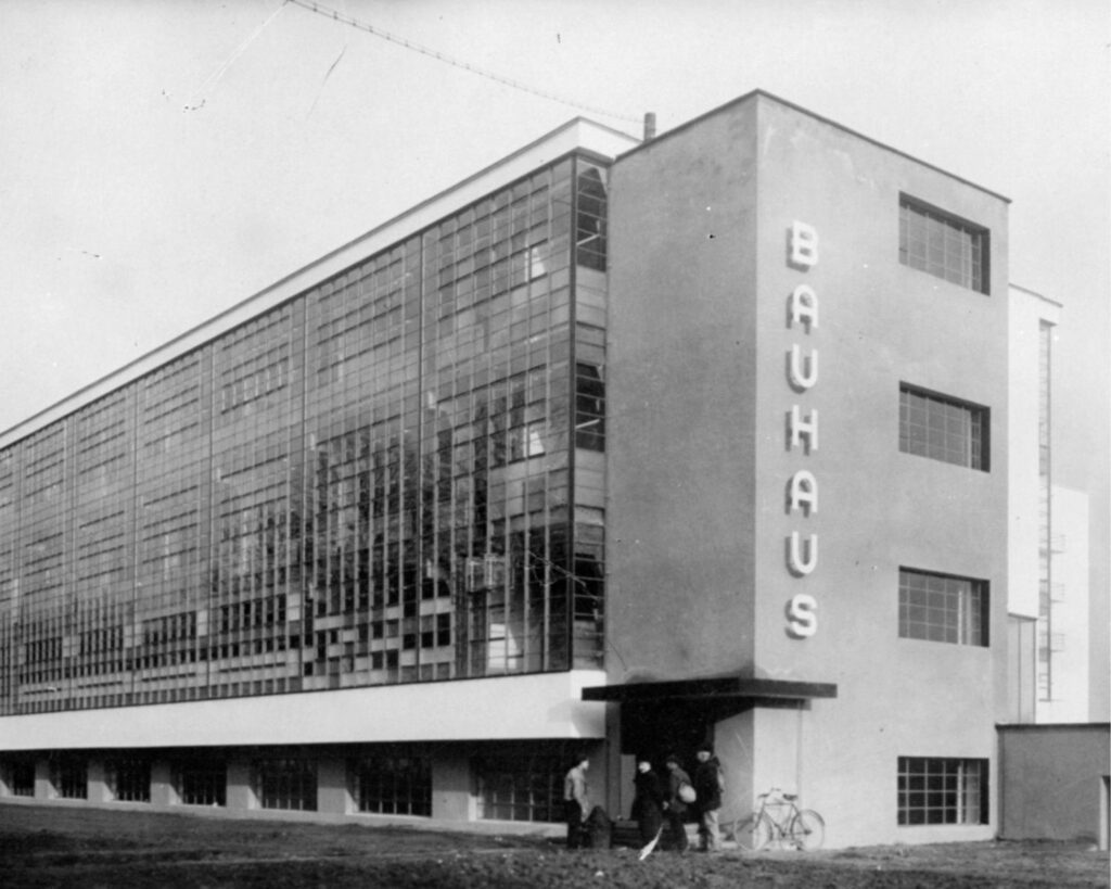 Historische Schwarzweißaufnahme des Bauhaus-Gebäudes in Dessau mit verglaster Werkstattfassade und vertikalem Schriftzug „Bauhaus“.