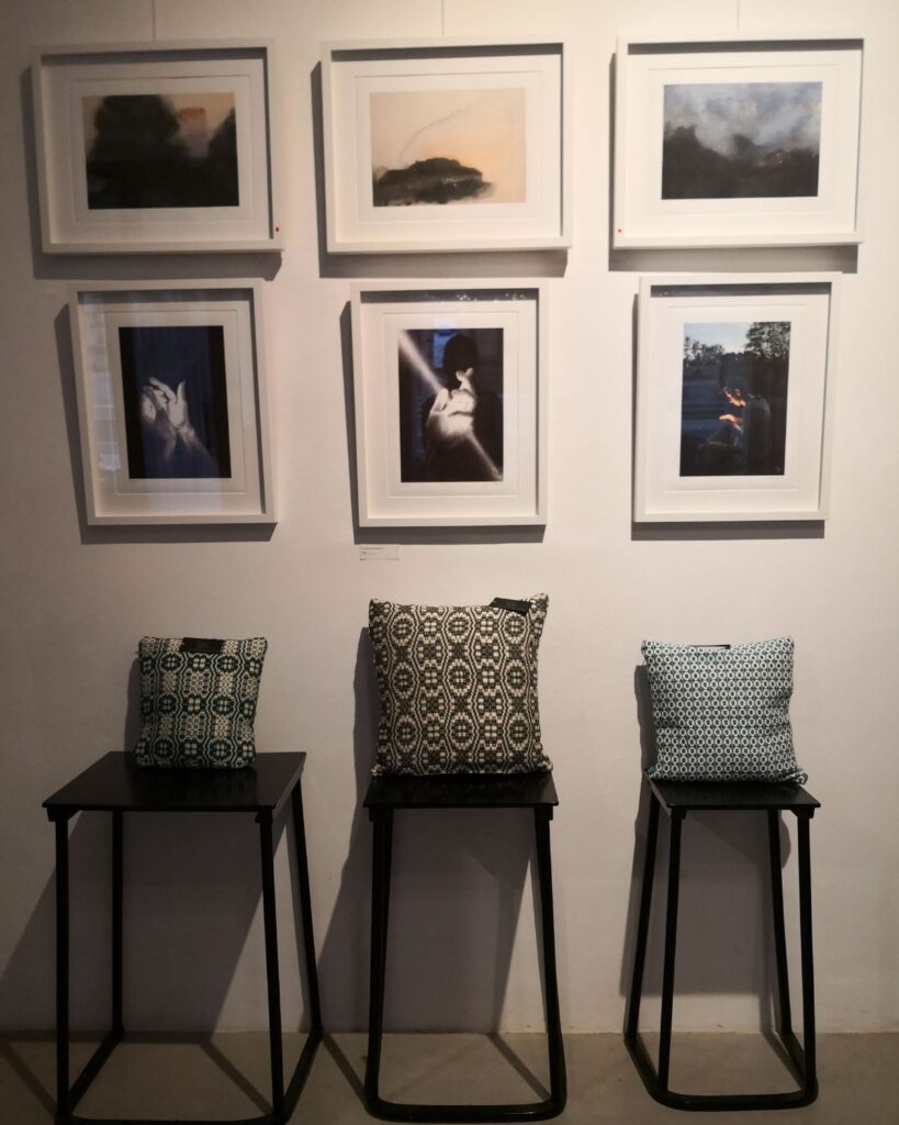 Handgewebte Kissen mit Ornamentvariationen auf Podesten unter gerahmten Kunstwerken in Graz 2025.