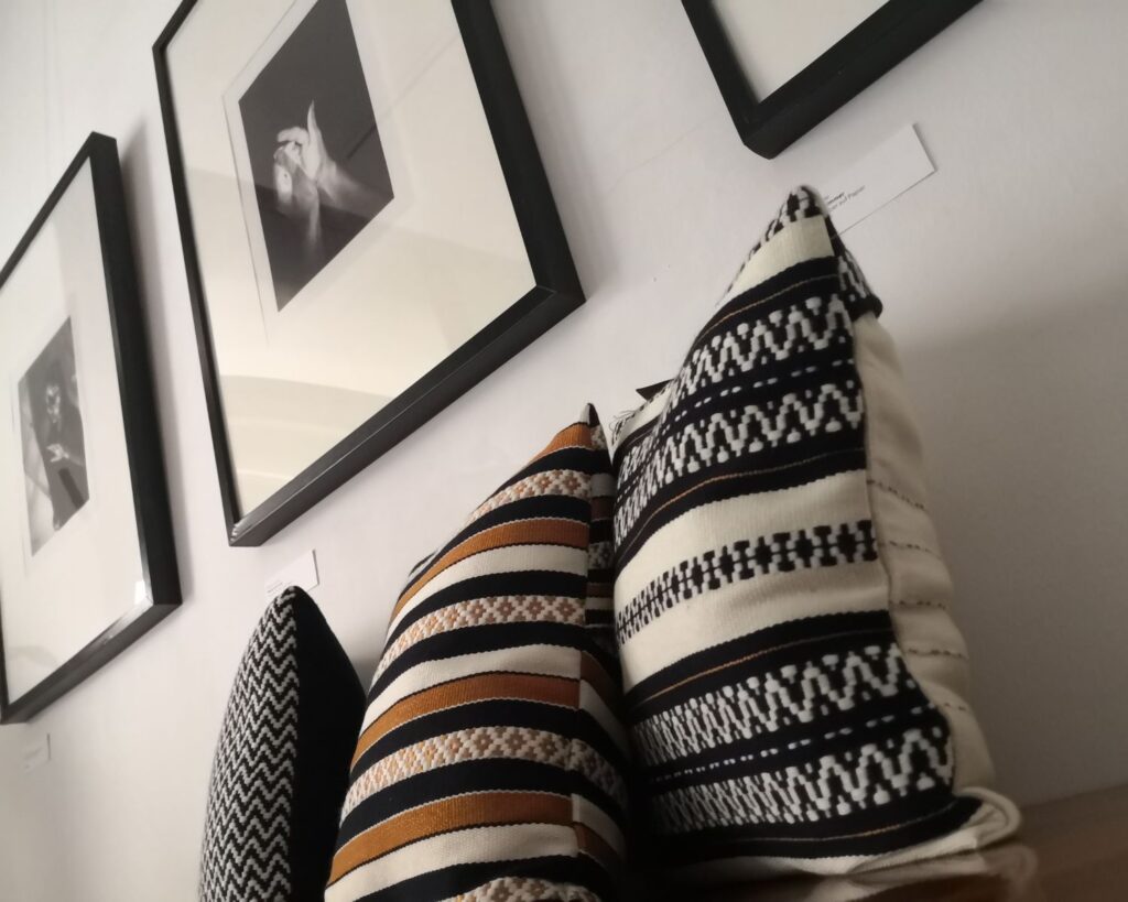 Handgewebte Kissen von Wiedengold aus Naturmaterial vor gerahmten Zeichnungen in Ausstellung in Graz 2025.