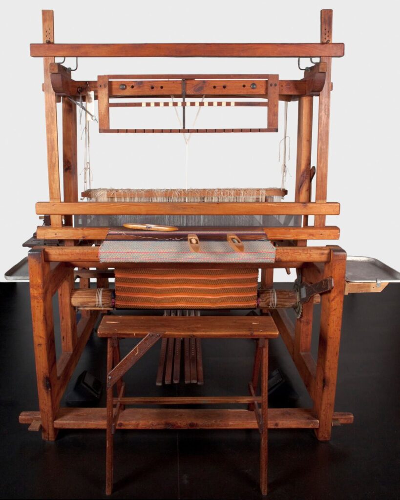 Holzwebstuhl mit eingespanntem Gewebe, Trittmechanik und Schiffchen, frontal aufgenommen.