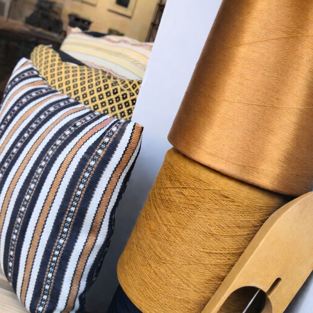 Handgewebte Kissen mit Ornamentik in Ocker, Blau und Weiß neben Garnkonen und einem Weberschiffchen.