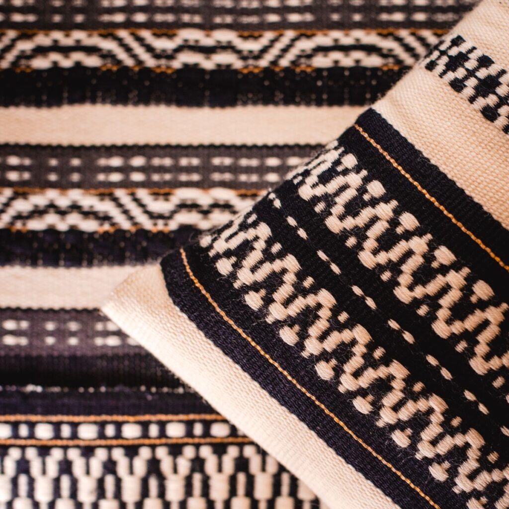 Handgewebtes Kissen und Webproben von Wiedengold mit ornamentaler Struktur und sichtbarer Fadenführung.